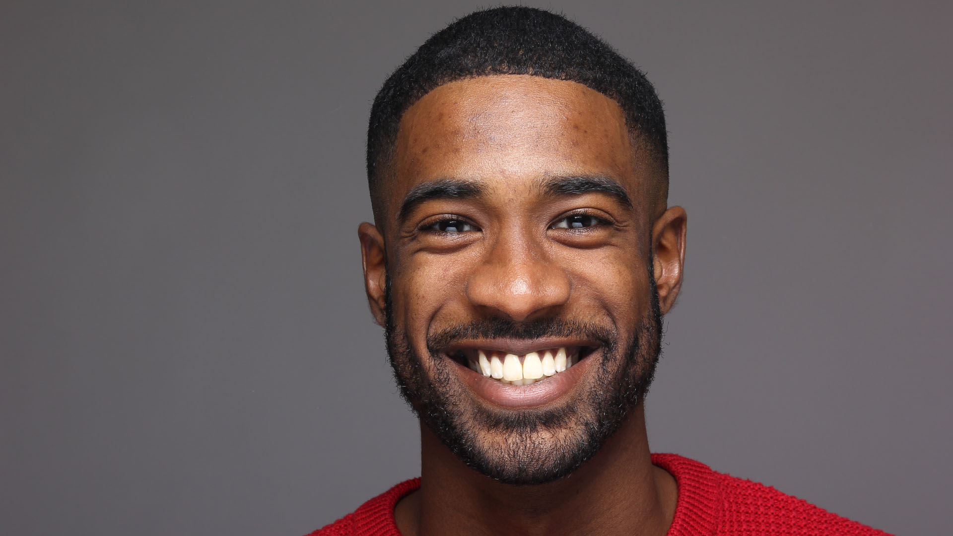 A young Black man with a short beard wearing a red jumper, smiling broadly against a grey studio background.